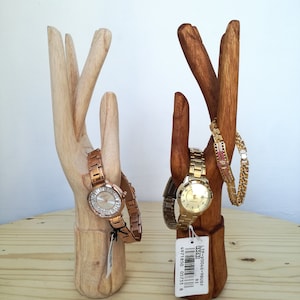 Puede incluir: Dos soportes de madera en forma de mano para relojes y pulseras. El soporte de la izquierda sostiene un reloj de oro rosa y el soporte de la derecha sostiene un reloj de oro y una pulsera de oro. El soporte de la derecha tiene una etiqueta de precio que dice "LTP-W004G-9BUDF 33523".