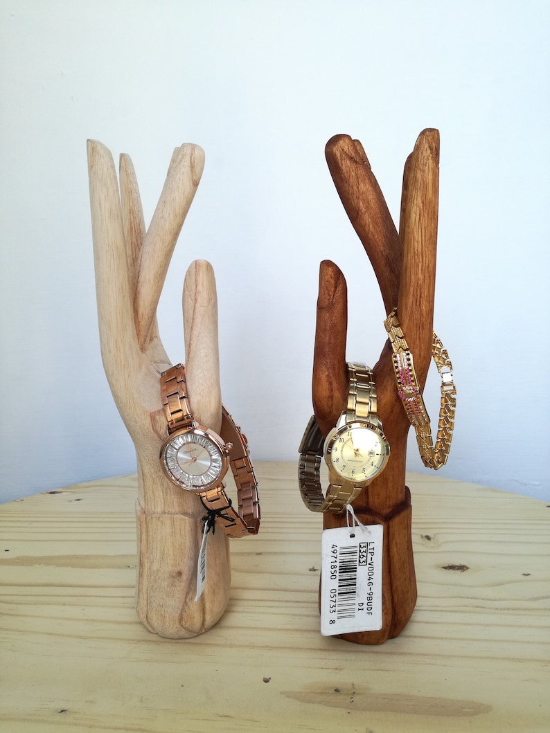 Puede incluir: Dos soportes de madera en forma de mano para relojes y pulseras. El soporte de la izquierda sostiene un reloj de oro rosa y el soporte de la derecha sostiene un reloj de oro y una pulsera de oro. El soporte de la derecha tiene una etiqueta de precio que dice "LTP-W004G-9BUDF 33523".