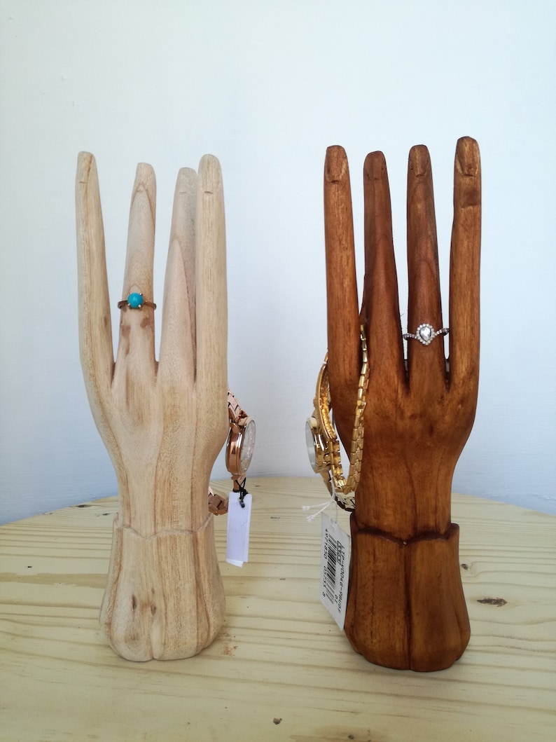 Puede incluir: Dos expositores de joyer&iacute;a en forma de mano de madera. Uno es de color madera natural y el otro es de color madera marr&oacute;n oscuro. Ambas manos tienen anillos y relojes.