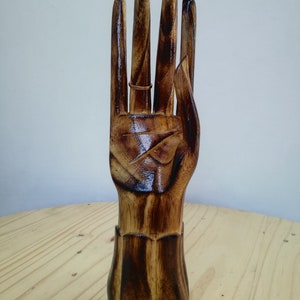 K&ouml;nnte beinhalten: Ein h&ouml;lzerner Ringhalter in Form einer Hand, dunkelbraun lackiert. Ein Ring wird an einem der Finger pr&auml;sentiert. Die Holzmaserung ist sichtbar, und die Basis ist heller braun. Der Ringhalter steht auf einer Holzoberfl&auml;che.
