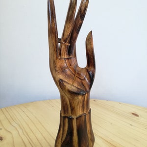 K&ouml;nnte beinhalten: Ein h&ouml;lzerner Ringhalter in Form einer Hand, dunkelbraun lackiert. Ein kleiner Ring wird an einem Finger pr&auml;sentiert. Der Sockel hat ein geschnitztes Design. Die Holzmaserung ist sichtbar. Der Halter ist aus Holz.