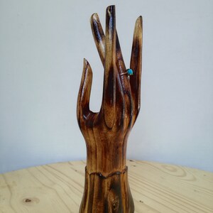 K&ouml;nnte beinhalten: Ein handf&ouml;rmiger Ringhalter aus Holz mit dunkelbraunem Finish. Ein goldfarbener Ring mit einem kleinen, runden, t&uuml;rkisfarbenen Stein befindet sich am Mittelfinger. Der Ringhalter steht auf einer hellbraunen Holzoberfl&auml;che.