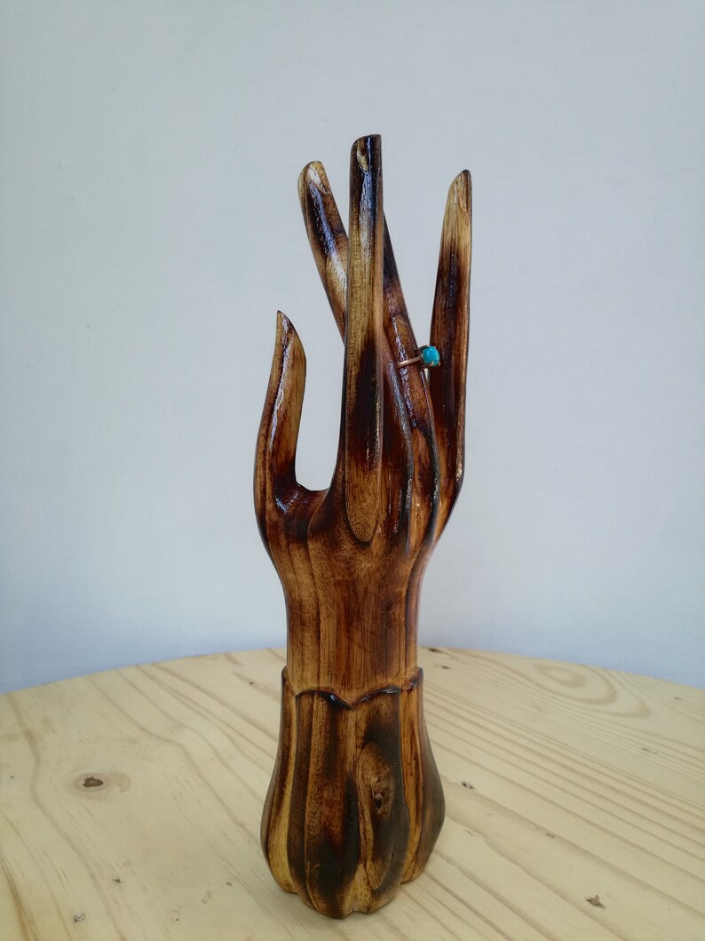 K&ouml;nnte beinhalten: Ein handf&ouml;rmiger Ringhalter aus Holz mit dunkelbraunem Finish. Ein goldfarbener Ring mit einem kleinen, runden, t&uuml;rkisfarbenen Stein befindet sich am Mittelfinger. Der Ringhalter steht auf einer hellbraunen Holzoberfl&auml;che.