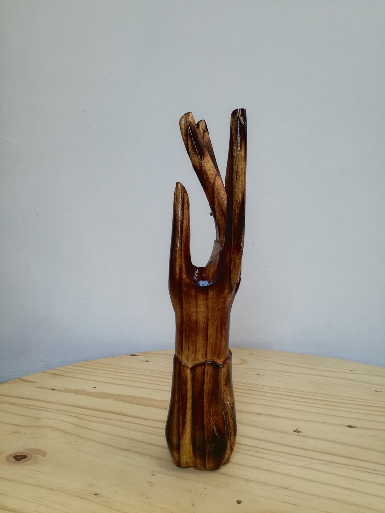 K&ouml;nnte beinhalten: Eine Holzskulptur einer Hand mit nach oben ausgestreckten Fingern. Die Skulptur besteht aus dunkelbraunem Holz mit gl&auml;nzender Oberfl&auml;che. Die Hand ist mit detaillierten Linien und Texturen geschnitzt, vor hellem Hintergrund.