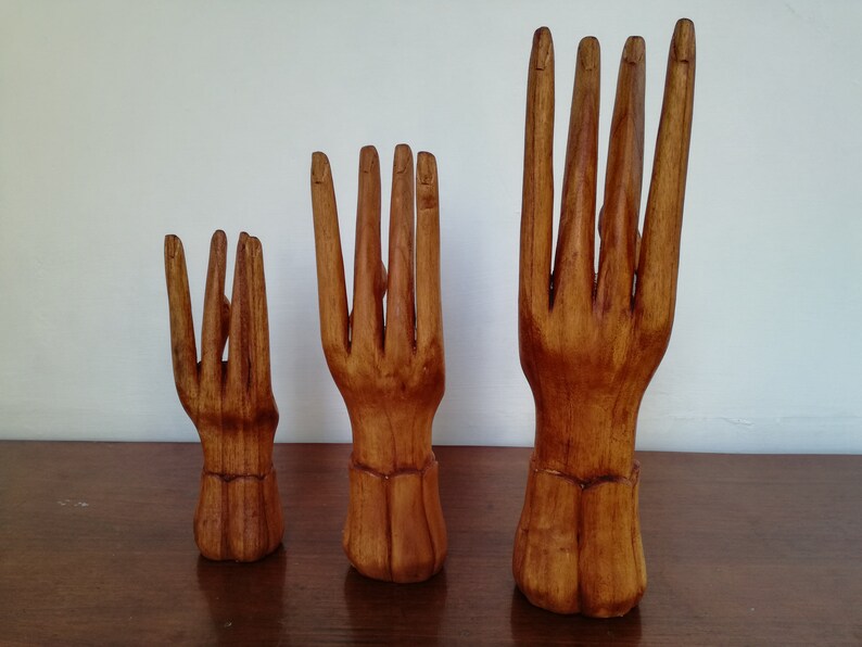Puede incluir: Tres soportes de anillos de madera con forma de mano en diferentes tama&ntilde;os. Las manos est&aacute;n talladas con detalles realistas y tienen un acabado de madera natural.