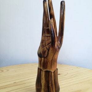 K&ouml;nnte beinhalten: Ein handf&ouml;rmiger Ringhalter aus Holz mit warmbraunem Finish. Die Finger sind ausgestreckt, und ein goldener Ring wird an einem der Finger pr&auml;sentiert. Der Sockel des Halters ist mit einem dekorativen Muster versehen.