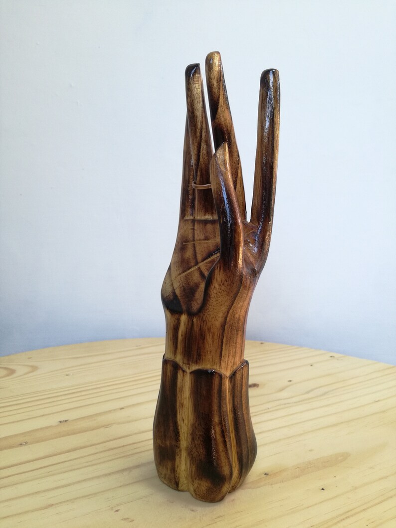 K&ouml;nnte beinhalten: Ein handf&ouml;rmiger Ringhalter aus Holz mit warmbraunem Finish. Die Finger sind ausgestreckt, und ein goldener Ring wird an einem der Finger pr&auml;sentiert. Der Sockel des Halters ist mit einem dekorativen Muster versehen.