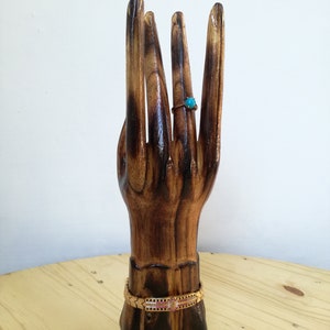 K&ouml;nnte beinhalten: Ein handf&ouml;rmiger Schmuckst&auml;nder aus Holz mit einem Ring und einem Armband. Der Ring hat einen kleinen, runden, t&uuml;rkisfarbenen Stein. Das Armband ist goldfarben mit rosa und schwarzen Akzenten. Der St&auml;nder ist aus dunkelbraunem Holz.