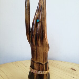 K&ouml;nnte beinhalten: Ein handf&ouml;rmiger Schmuckst&auml;nder aus Holz mit einem goldenen Armband und einem Ring mit einem t&uuml;rkisfarbenen Stein. Der St&auml;nder ist aus dunkelbraunem Holz mit gl&auml;nzendem Finish gefertigt und steht auf einer hellbraunen Holzoberfl&auml;che.
