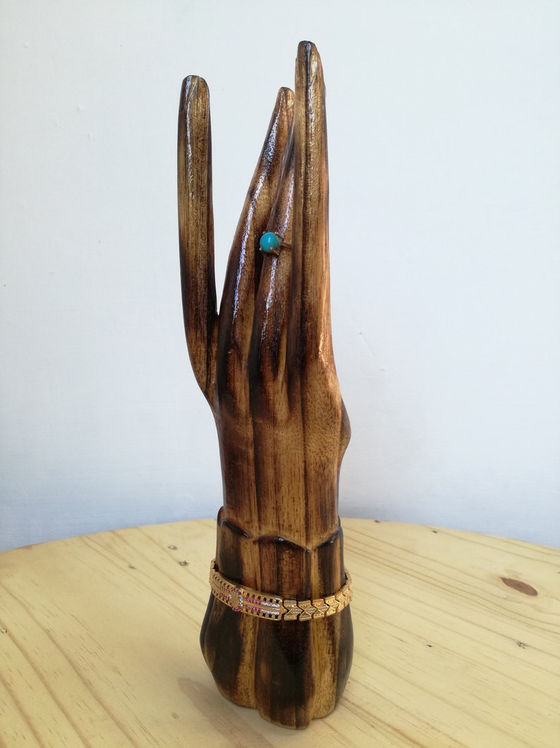K&ouml;nnte beinhalten: Ein handf&ouml;rmiger Schmuckst&auml;nder aus Holz mit einem goldenen Armband und einem Ring mit einem t&uuml;rkisfarbenen Stein. Der St&auml;nder ist aus dunkelbraunem Holz mit gl&auml;nzendem Finish gefertigt und steht auf einer hellbraunen Holzoberfl&auml;che.
