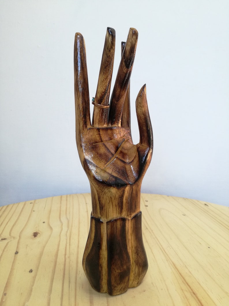 K&ouml;nnte beinhalten: Ein h&ouml;lzerner, handf&ouml;rmiger Ringhalter mit dunkelbraunem Finish. Der Ringhalter ist aus geschnitztem Holz gefertigt und hat einen Ring am Mittelfinger. Der Ringhalter steht auf einer hellbraunen Holzoberfl&auml;che.