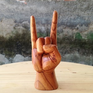 May include: A hand-carved wooden sculpture depicting a hand making the rock and roll sign. The object features a warm brown wood grain with visible natural textures, showing the index and pinky fingers extended upward while the thumb holds the middle fingers.