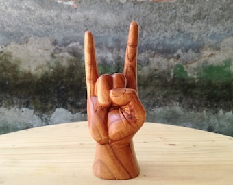 May include: A hand-carved wooden sculpture depicting a hand making the rock and roll sign. The object features a warm brown wood grain with visible natural textures, showing the index and pinky fingers extended upward while the thumb holds the middle fingers.