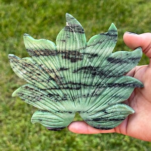 May include: A carved green stone sculpture shaped like a fan of leaves with dark brown and black horizontal striations. The object features a series of radiating grooves that define the individual leaf segments, creating a textured surface.