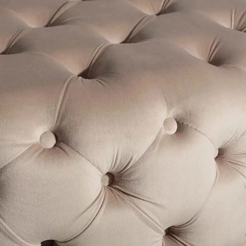 K&ouml;nnte beinhalten: Nahaufnahme eines beigefarbenen, gesteppten Ottomans mit Knopfdetails. Der Stoff ist weich und pl&uuml;schig.