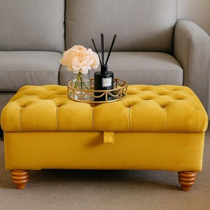 May include: A mustard yellow velvet ottoman with wooden legs. A gold-coloured tray holds a vase of flowers and a black diffuser bottle. The ottoman is in front of a grey sofa.