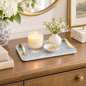 May include: A rectangular serving tray with a blue and white floral pattern and gold-toned metal handles. On the tray sits a lit white candle in a glass jar, a small white ceramic vase with white flowers, and a small scalloped white dish.