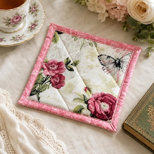 May include: A square quilted fabric coaster featuring a floral pattern with deep pink roses and a black and white butterfly on a cream background. The coaster is finished with a bright pink floral border and diagonal stitching across the surface.