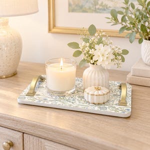 May include: A rectangular serving tray with a blue and green floral pattern and gold-toned metal handles. The tray holds a clear glass candle holder with a lit white candle, a small white ribbed vase with white flowers, and a small white lidded box.
