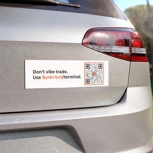 May include: A white rectangular bumper sticker with the text "Don't vibe trade. Use Bankr.bot/terminal." and a QR code with an orange centre, attached to the rear of a silver car.