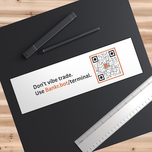 May include: A white sticker with the text "Don't vibe trade. Use Bankr.bot/terminal." and a QR code with an orange border. Two black pens and a ruler are also visible.