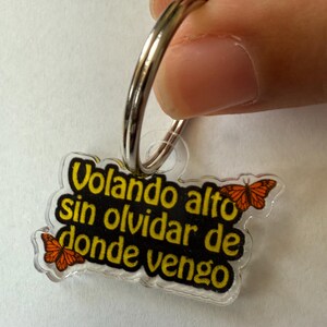 May include: A clear acrylic keychain with a silver metal ring. The keychain features the Spanish text "Volando alto sin olvidar de donde vengo" in yellow lettering on a black background, with orange butterfly graphics.