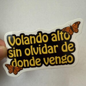 May include: A white sticker with the Spanish text "Volando alto sin olvidar de donde vengo" in yellow letters with a black outline. Two orange butterflies are on the sticker.