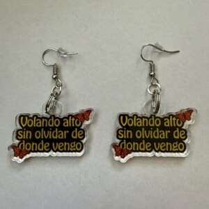 May include: A pair of dangle earrings with a clear, rectangular charm. The charm has the text "Volando alto sin olvidar de donde vengo" in black and yellow, with orange butterfly accents. The earrings have silver-colored hooks.