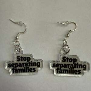 May include: A pair of clear acrylic earrings with silver-colored hooks. Each earring has the phrase "Stop separating families" in black text. The earrings are designed to be worn and are displayed against a white background.