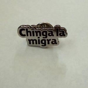 May include: A silver-colored pin with the words "Chinga la migra" in black lettering. The pin has a rounded, cloud-like shape and a pin backing.