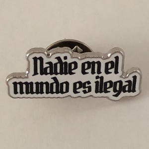 May include: A silver-colored pin with the phrase "Nadie en el mundo es ilegal" in black lettering with a white border. The pin has a classic pin back.