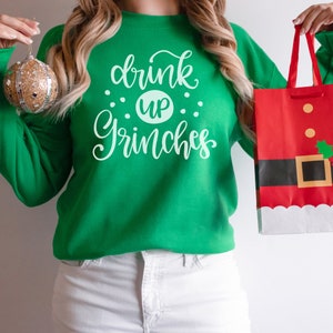 May include: A woman wearing a green sweatshirt with the text "drink up Grinches" in white. She is holding a red and white gift bag with a Santa Claus design.