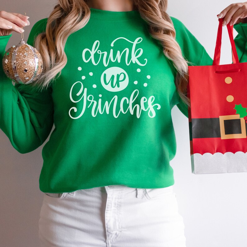 May include: A woman wearing a green sweatshirt with the text "drink up Grinches" in white. She is holding a red and white gift bag with a Santa Claus design.