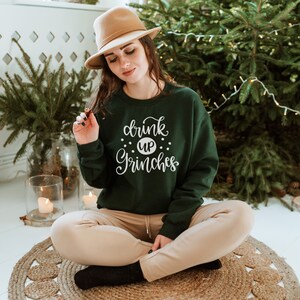 May include: A woman wearing a green sweatshirt with the text "drink up Grinches" in white lettering. She is sitting on a woven rug with a Christmas tree in the background.