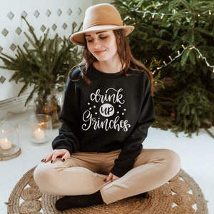 May include: A black sweatshirt with the text "drink up Grinches" in white lettering. The sweatshirt is being worn by a person sitting on a woven rug.