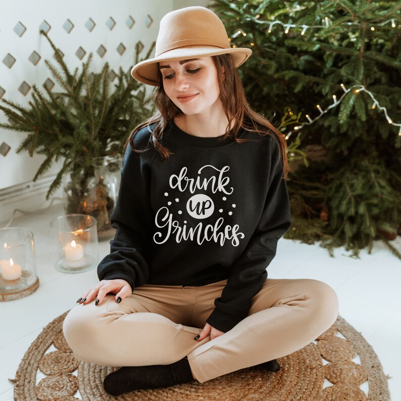 May include: A black sweatshirt with the text "drink up Grinches" in white lettering. The sweatshirt is being worn by a person sitting on a woven rug.