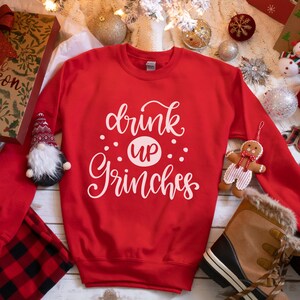 May include: Red Christmas sweatshirt with white text that reads "drink up Grinches". The sweatshirt is on a white wooden surface with Christmas decorations.