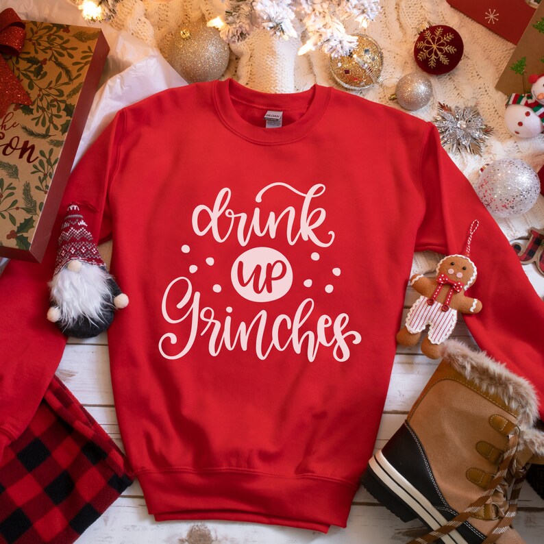 May include: Red Christmas sweatshirt with white text that reads "drink up Grinches". The sweatshirt is on a white wooden surface with Christmas decorations.