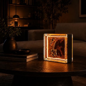 Op de afbeelding: Een vierkante glazen bloklamp met een warme, amberkleurige interne lichtbron die een vloeiend, golvend patroon in het transparante glas verlicht. De lamp heeft een donker, solide frame aan de zijkant en geeft zacht licht.