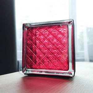 Puede incluir: Un bloque de vidrio cuadrado de tono rojo vibrante con un patr&oacute;n de rejilla de diamantes en relieve en su superficie. El bloque presenta un marco de borde liso y transparente, con una construcci&oacute;n gruesa y s&oacute;lida que refleja la luz.