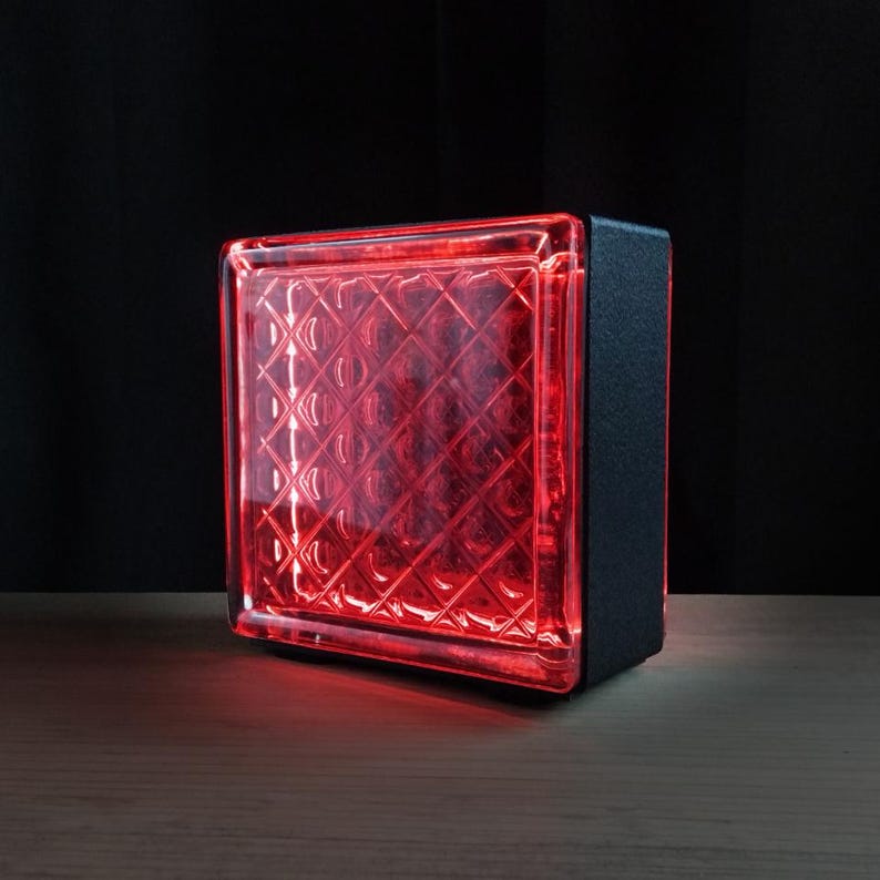Puede incluir: Una l&aacute;mpara de bloque de vidrio cuadrada que emite una luz roja vibrante. La cara frontal presenta un patr&oacute;n de rejilla de diamante en relieve, mientras que el lado y la parte posterior est&aacute;n encerrados en un marco negro s&oacute;lido.