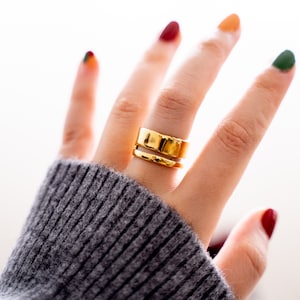 May include: A gold ring with a wide band and a thinner band on top. The ring is worn on the middle finger of a hand with colorful nail polish.