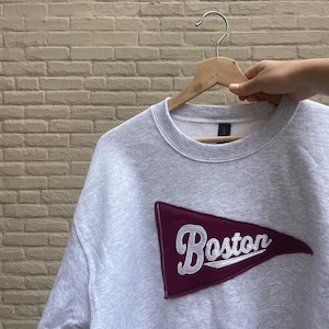 May include: A light gray sweatshirt with a maroon pennant patch featuring the word "Boston" in white script. The sweatshirt is hanging on a wooden hanger against a light brick wall.