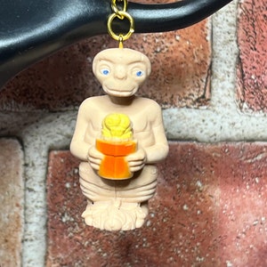 May include: A beige plastic keychain featuring a figurine of E.T. holding a small orange box with a yellow object inside. The keychain has a gold metal chain.