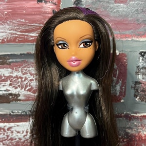 Puede incluir: Una muñeca con cabello castaño y reflejos morados. La muñeca tiene ojos marrones, labios rosados y un torso plateado. La muñeca está en un soporte negro.