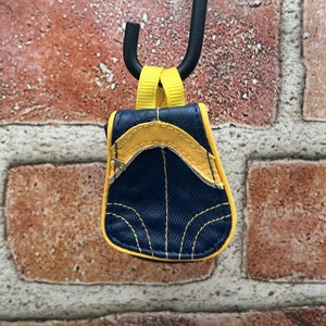 Puede incluir: Un pequeño charm de mochila azul y amarillo con cierre de cremallera. El charm está colgado de un gancho negro.
