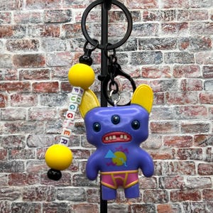 May include: A novelty keychain featuring a purple monster with three eyes, yellow ears, and a wide grin. The keychain includes a beaded lanyard spelling "FUGGLER". The monster wears pink and yellow underwear. The keychain is attached to a black metal ring.