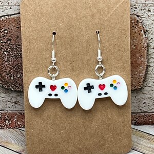Puede incluir: Un par de pendientes blancos de mando de videojuego con corazones rojos y botones de colores. Los pendientes están en una tarjeta marrón.