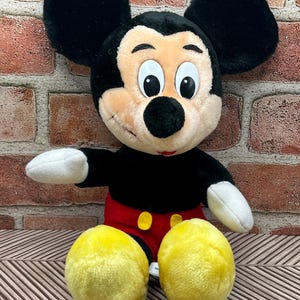 May include: A plush Mickey Mouse toy sitting with his legs crossed. He is wearing red shorts with yellow buttons and has large black ears and yellow shoes.