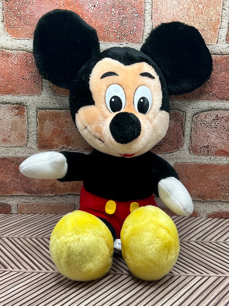 May include: A plush Mickey Mouse toy sitting with his legs crossed. He is wearing red shorts with yellow buttons and has large black ears and yellow shoes.